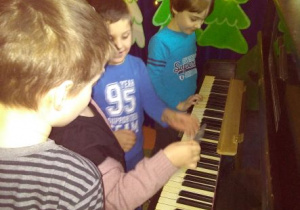 Dzieci stoją przed otwartą klawiaturą pianina. Dziewczynka trzyma odnalezioną kartę z zadaniem